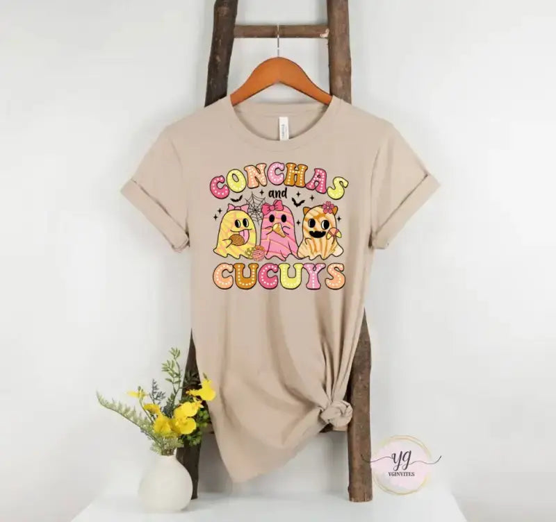 Beige t-shirt with colorful ’Conchas Cuguys’ cartoon dog design on the front.