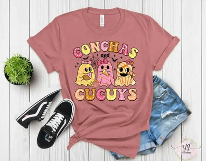 Mauve t-shirt featuring cartoon conchas (Mexican sweet bread) characters with the text ’CONCHAS AND CUGUYS’