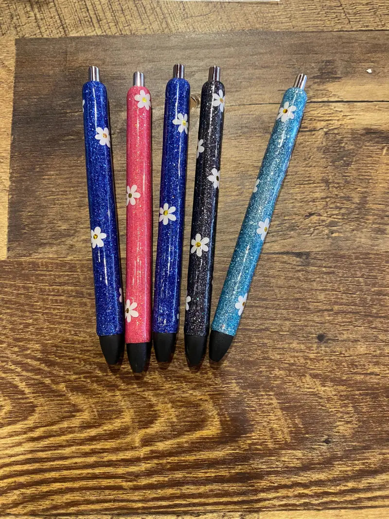 Five glittery pens with flower decorations in blue, pink, and black colors.