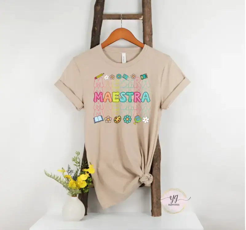 Beige t-shirt with colorful ’MAESTRA’ text and decorative elements displayed on a wooden hanger.