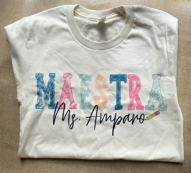 White t-shirt with ’MAESTRA Ms Amparo’ printed in colorful patterned letters.