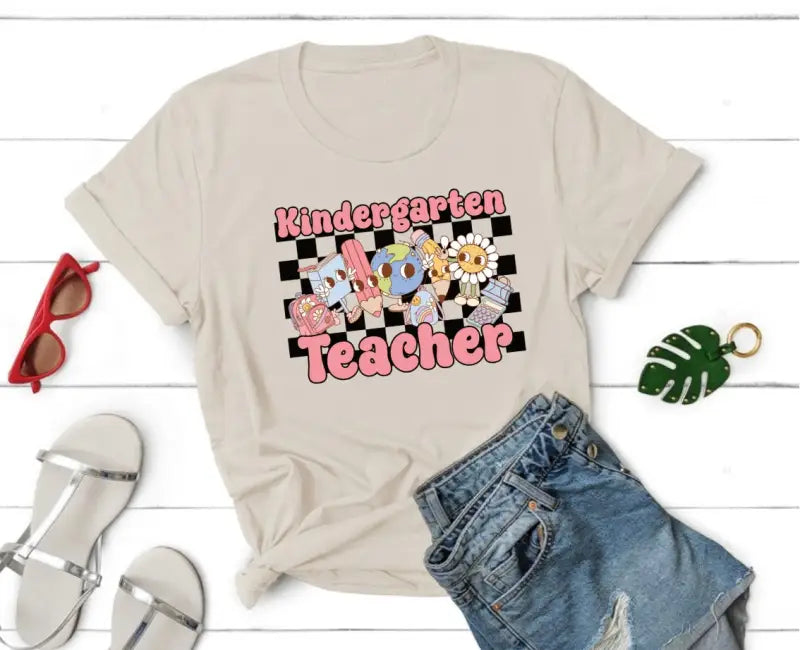 Beige t-shirt with ’Kindergarten Teacher’ text in pink and checkered pattern design.